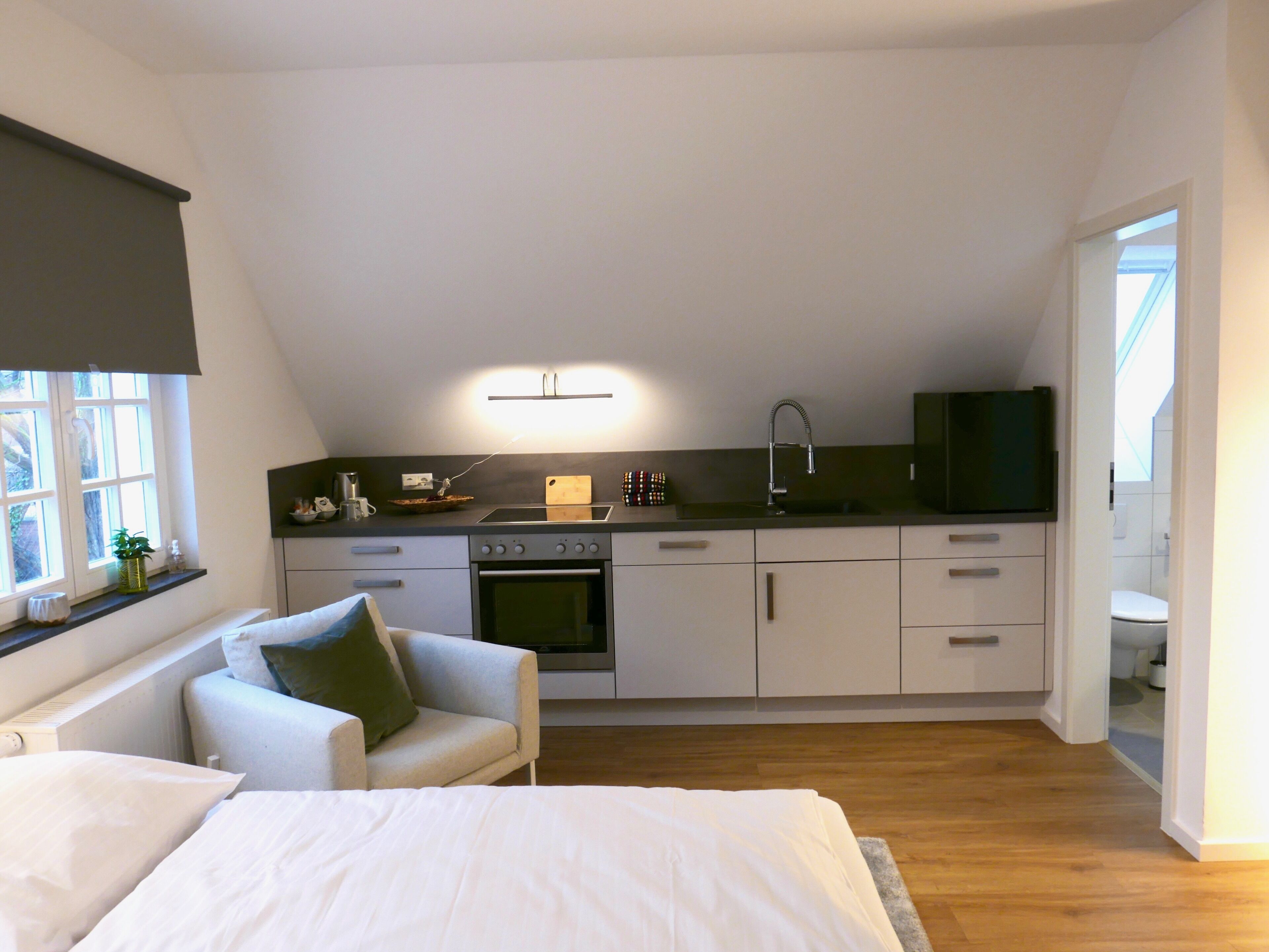A compact kitchen with a built-in oven, a sink, and a small countertop. There is a sofa and a bed in the adjacent room.