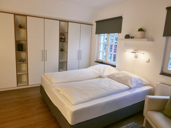 Un dormitorio con cama, armario, estante con plantas, ventana y radiador.