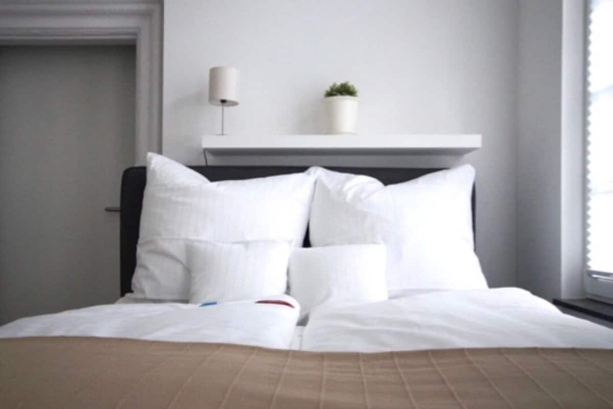 A neatly made bed with white bedding and a brown accent, a bedside lamp, a small potted plant on a white shelf, and a window with blinds.