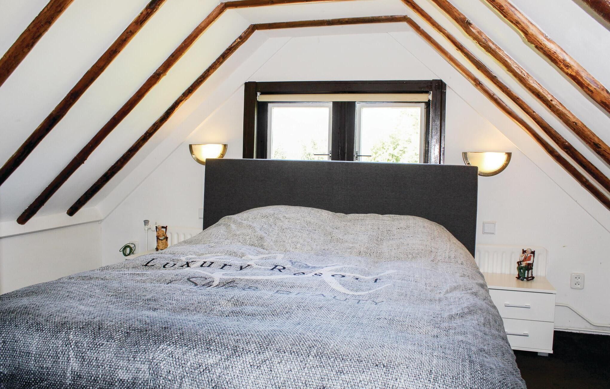 Un dormitorio con techo inclinado, una cama con cabecera gris, dos lámparas de pared, un ventanal y una mesita de noche con una figurita.