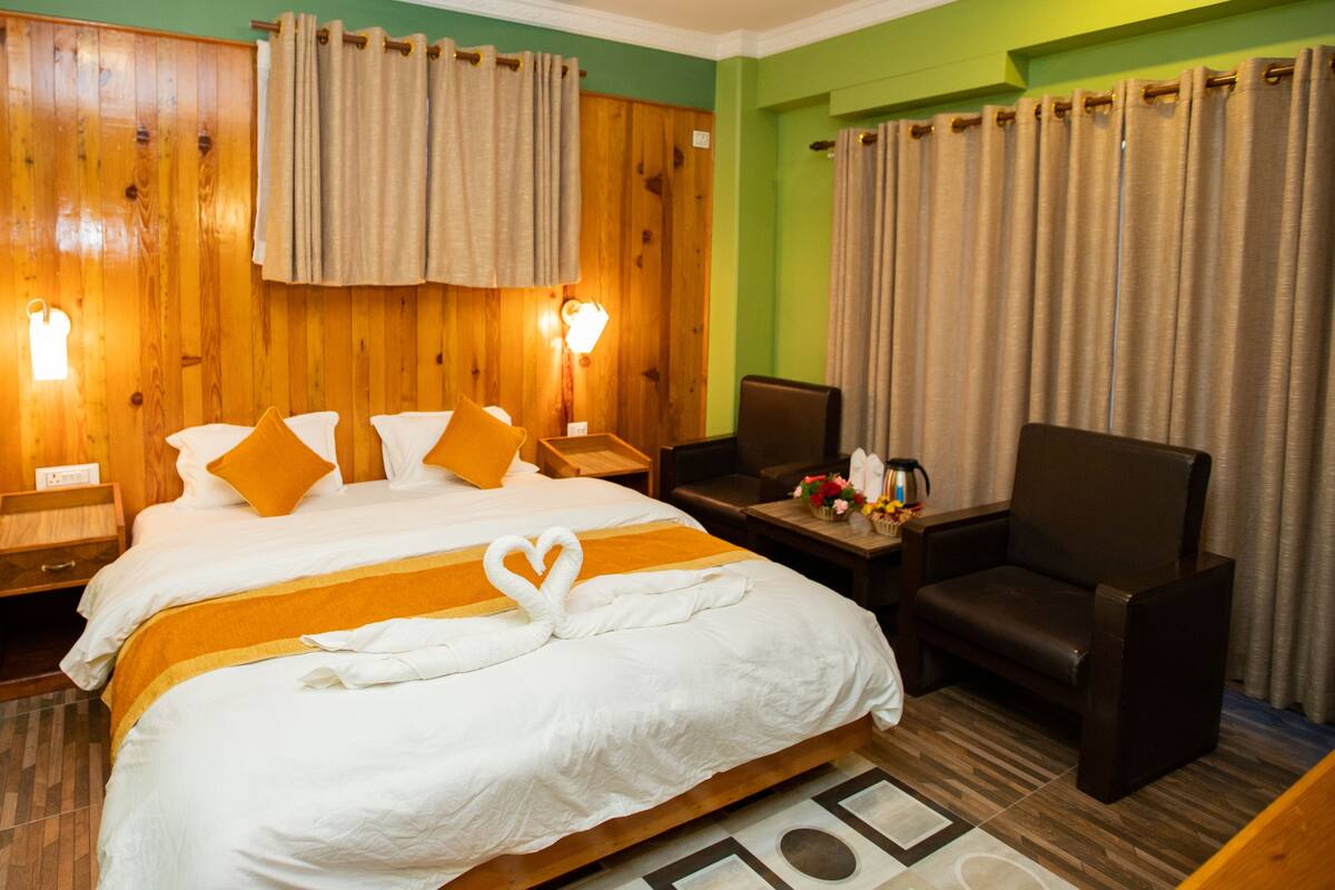 Une chambre d’hôtel avec une tête de lit en bois, un lit avec une literie blanche et orange, une serviette en forme de cœur posée sur le lit, un bureau avec une chaise et des rideaux aux fenêtres.