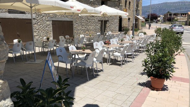 An outdoor cafe with white plastic chairs and tables, covered by white umbrellas, set against a stone building with a tiled roof. - Casa Puyuelo (La Fueva)