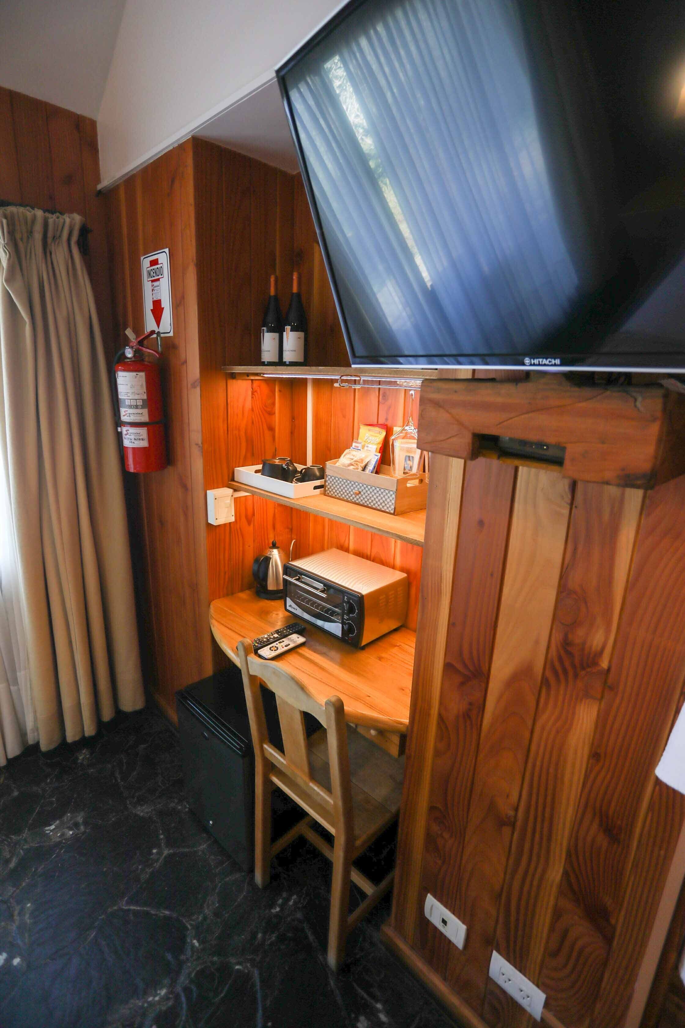A compact room with a television, wooden shelves, a fire extinguisher, and a small table with a chair.
