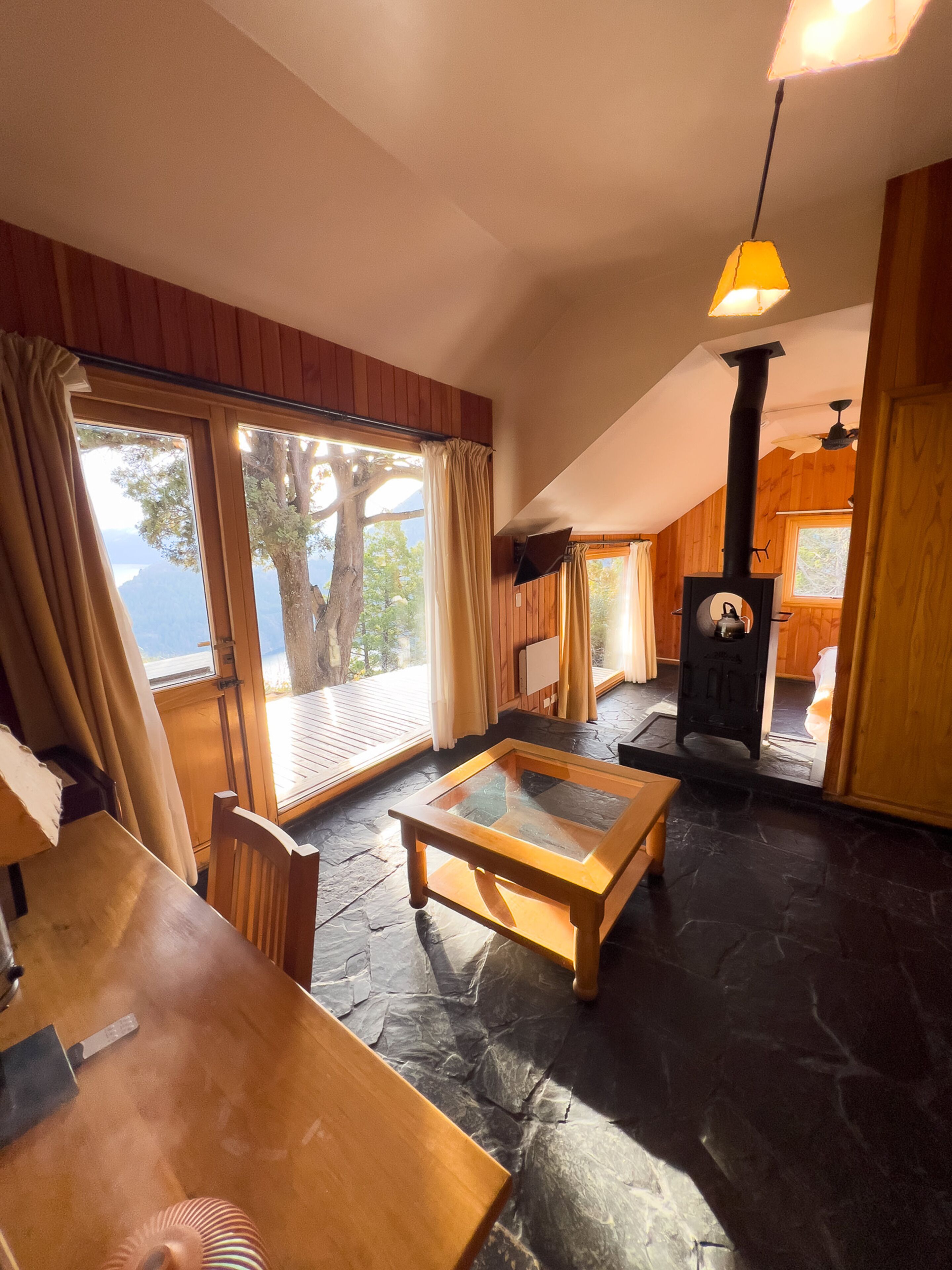 A cozy room with a large window, a wooden table, a stone floor, and a black stove.