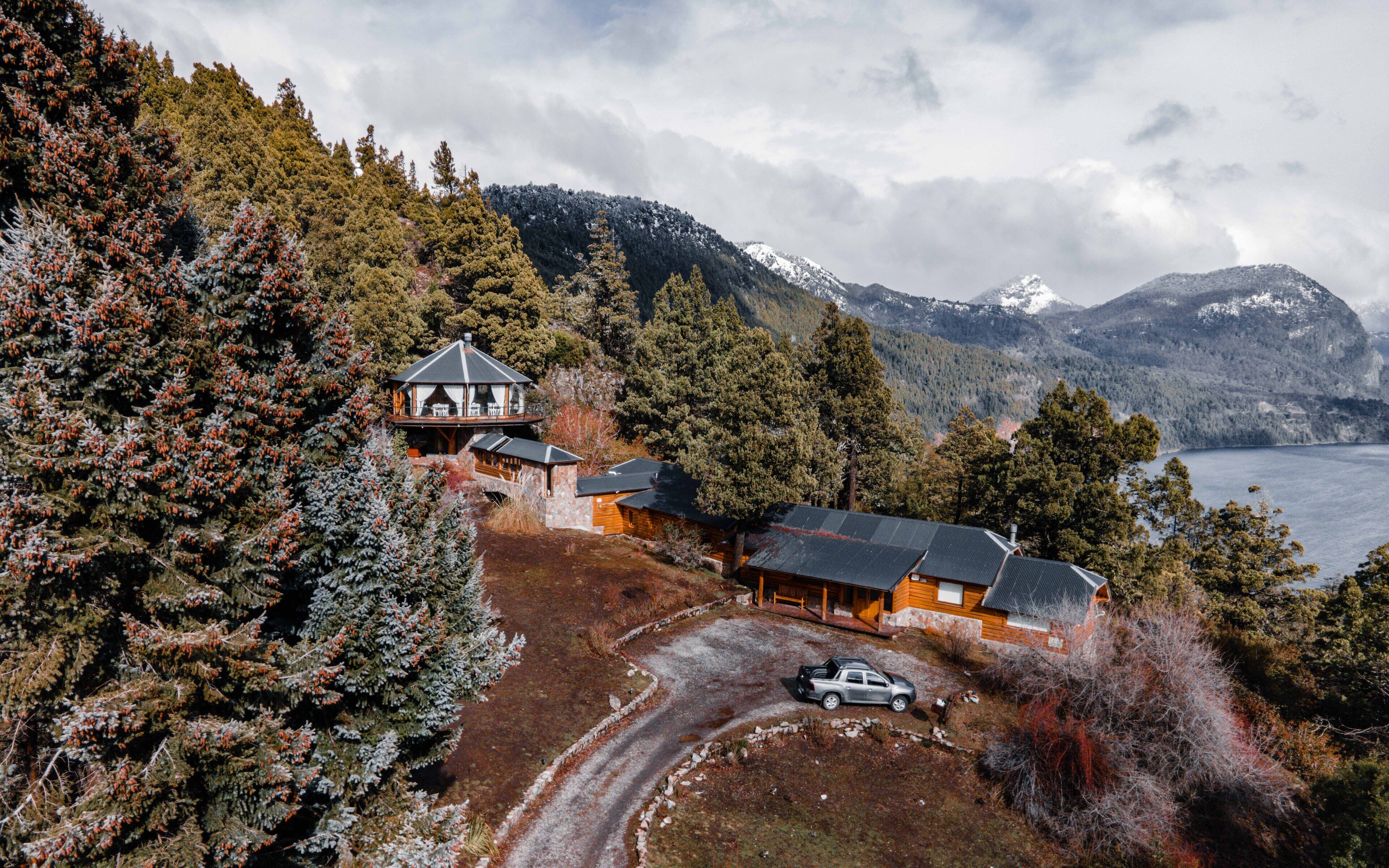 Eine Lodge mit großer Terrasse, mehreren Hütten und Blick auf See und Berge.