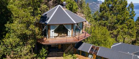 Eine Hütte mit einem Balkon, umgeben von dichtem Wald und Blick auf einen See und Berge.