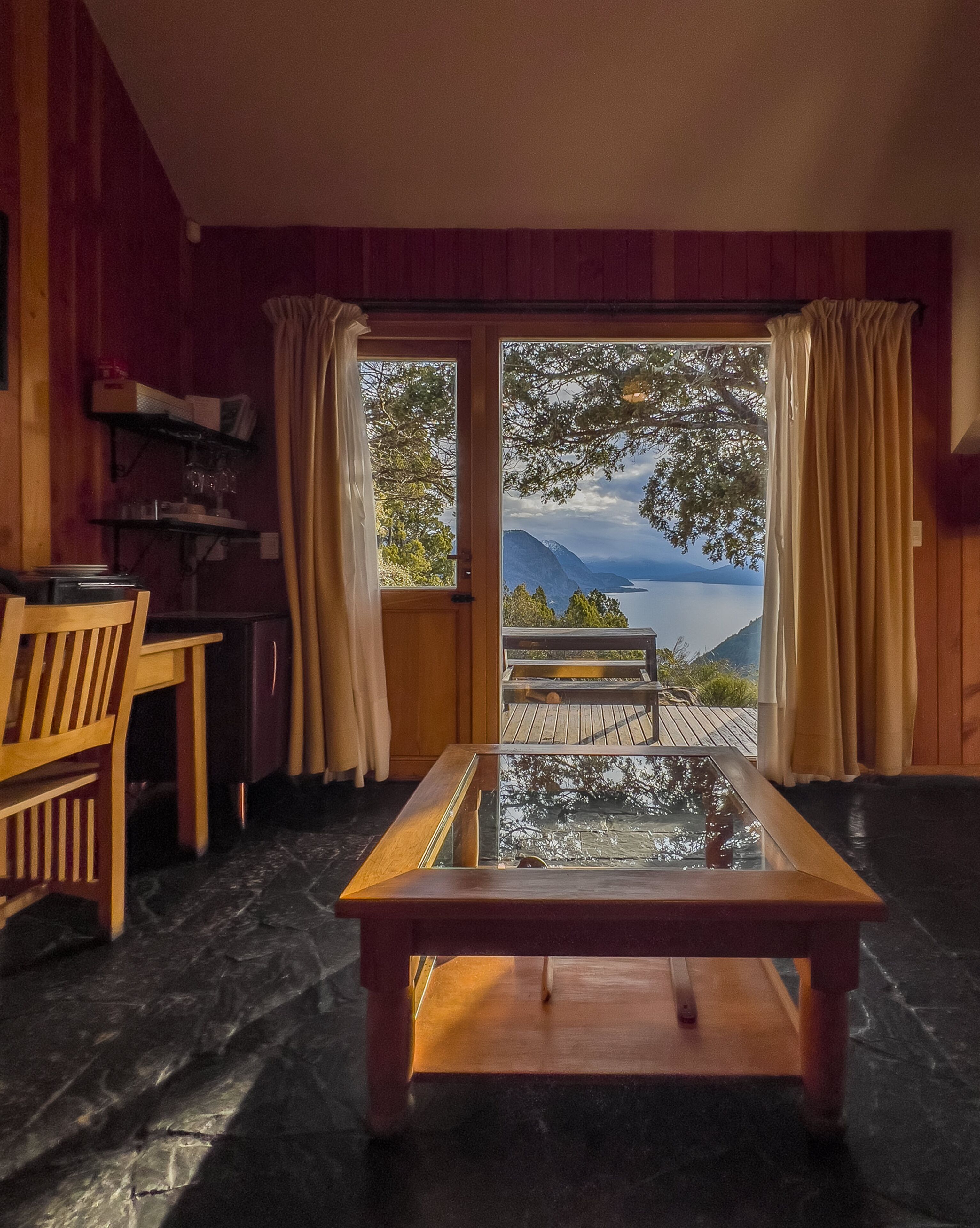 A room with a wooden table and chairs, a glass-top table, and a view of a lake and mountains through large windows.