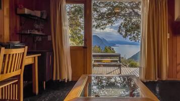 A room with a wooden table and chairs, a glass-top table, and a view of a lake and mountains through large windows.