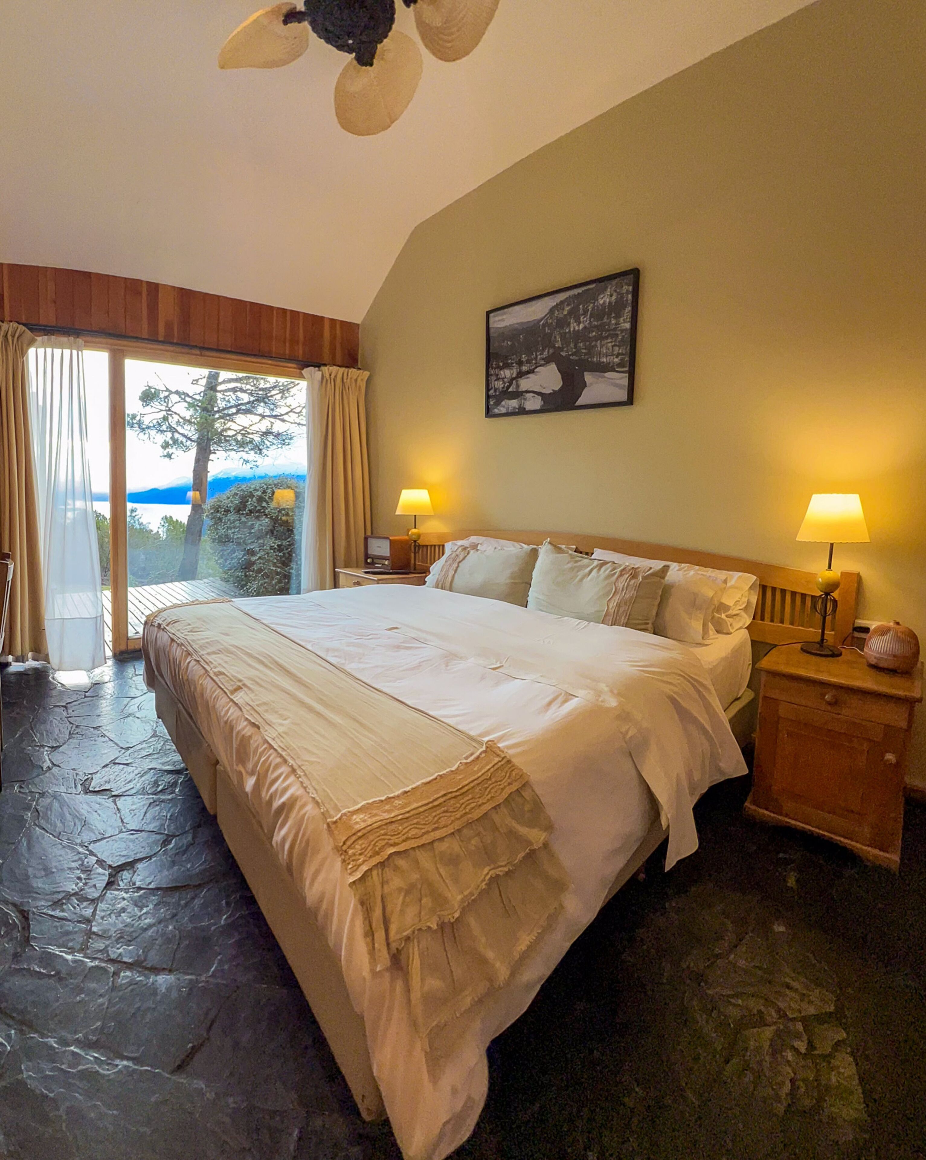 A bedroom with a large bed, two bedside tables with lamps, a painting on the wall, and a view of the outdoors.