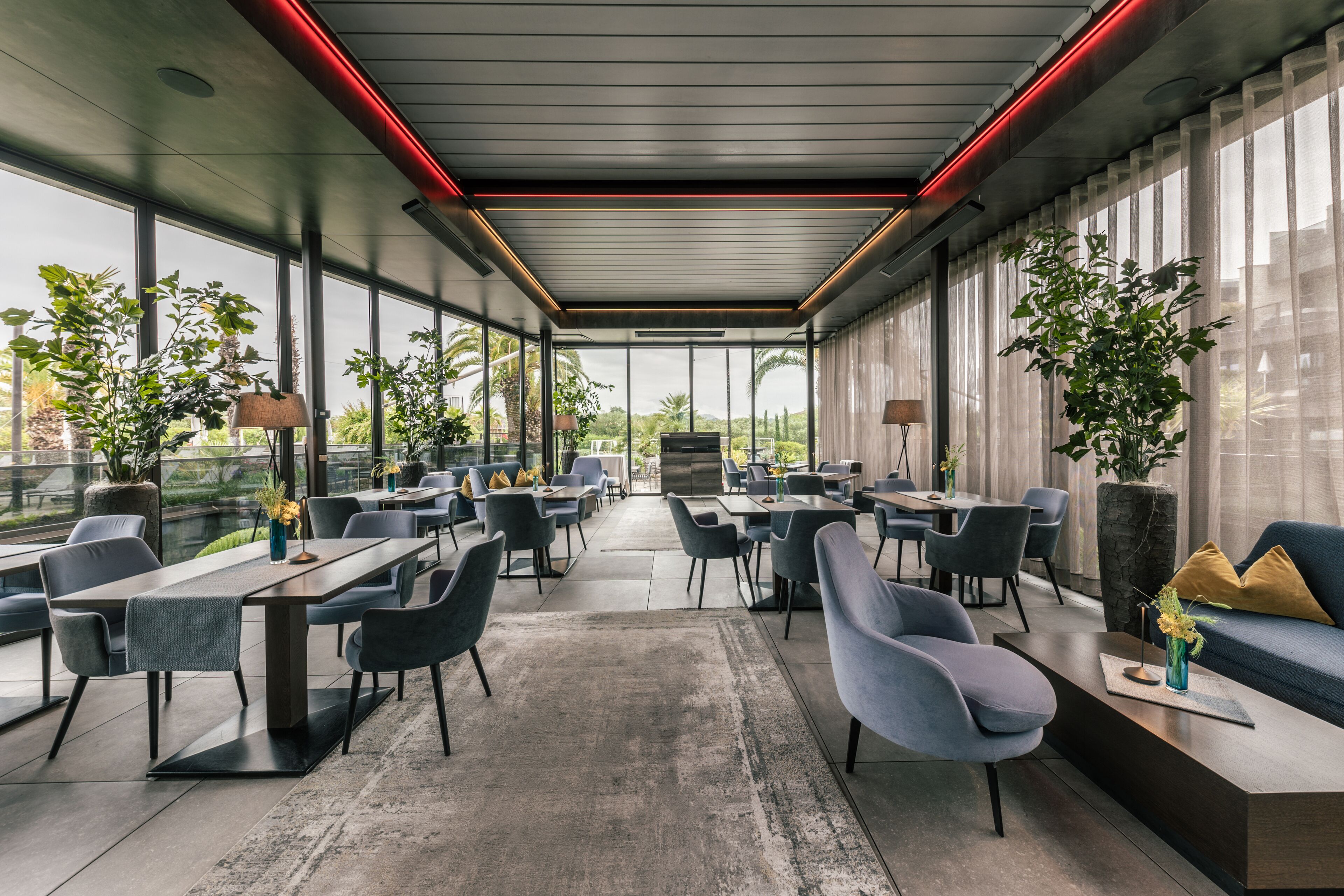 A modern indoor space with tables and chairs arranged for dining or meetings, large windows offering views of the outdoors, and potted plants adding greenery.