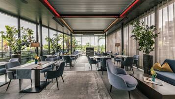 A modern indoor space with tables and chairs arranged for dining or meetings, large windows offering views of the outdoors, and potted plants adding greenery.