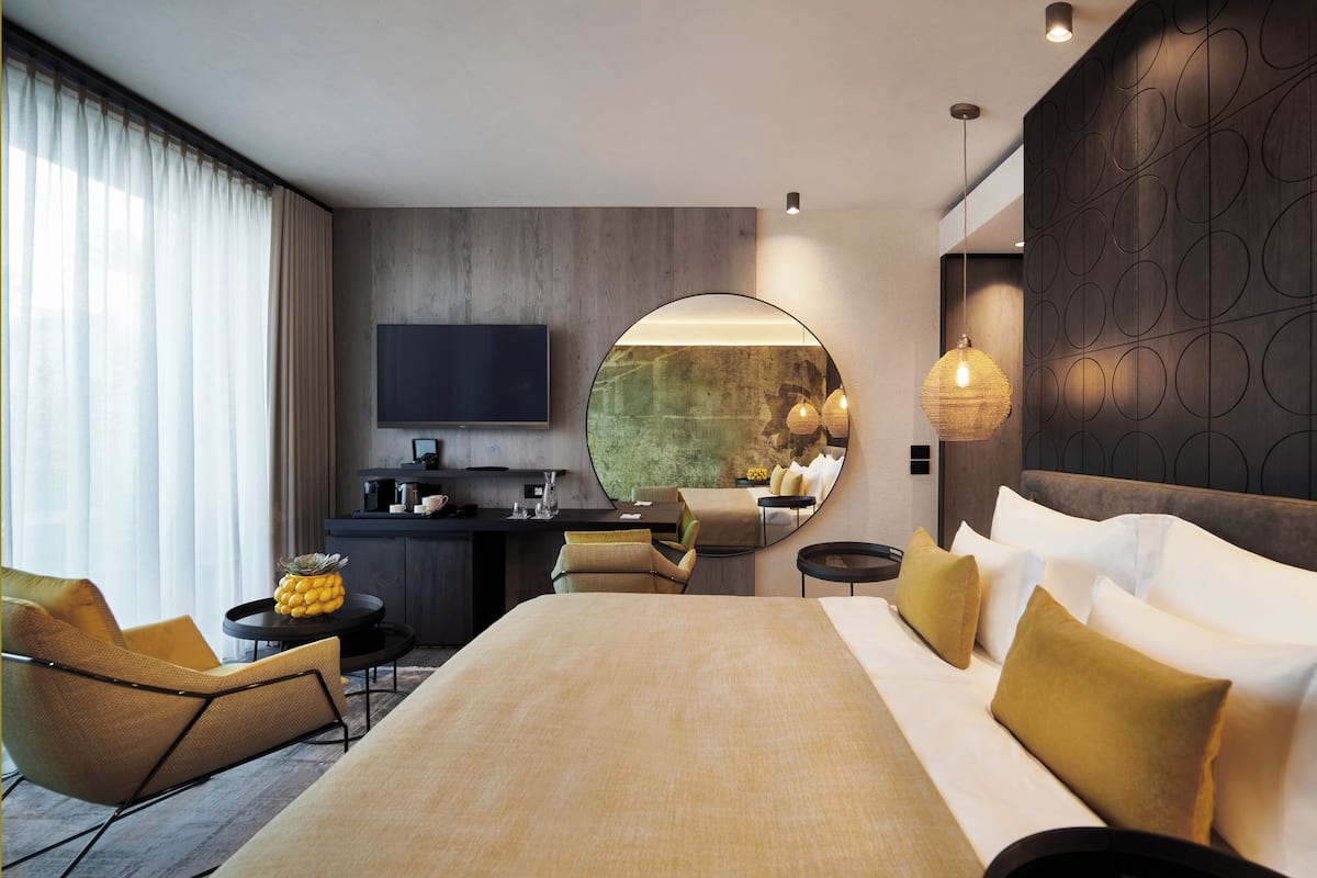 Una habitación de hotel moderna con una cama grande, un escritorio con televisor de pantalla plana, un espejo redondo y una zona de estar con sillas y una mesita.