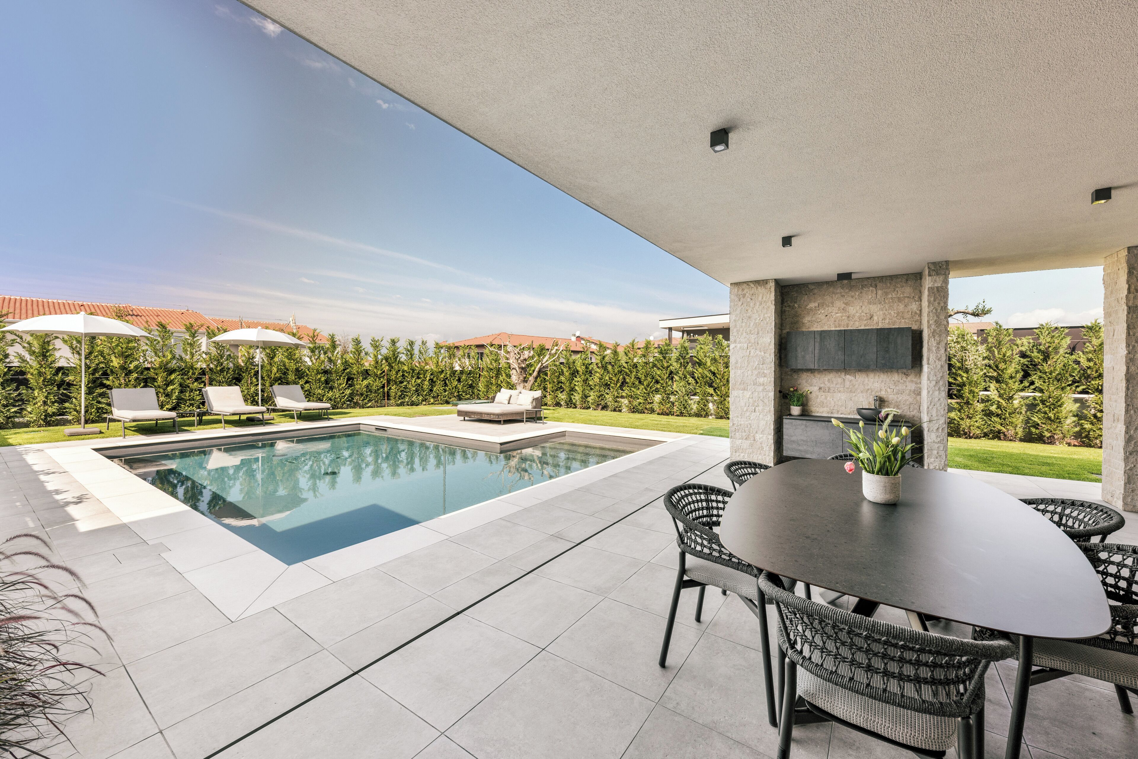 A modern outdoor pool area with a dining table and chairs, surrounded by greenery and a clear sky.