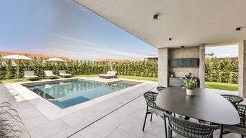 A modern outdoor pool area with a dining table and chairs, surrounded by greenery and a clear sky.
