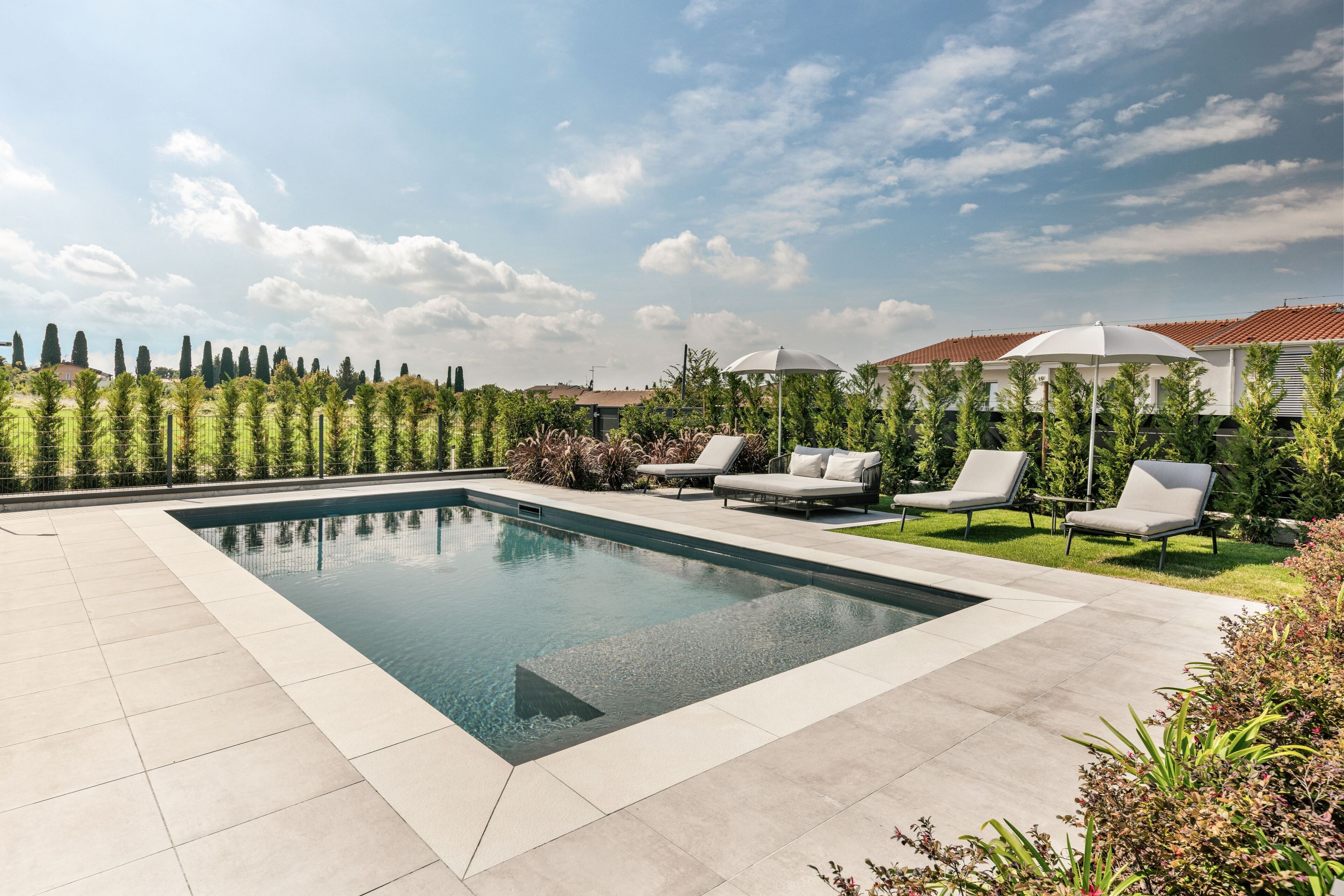 A rectangular swimming pool with a tiled deck, surrounded by lounge chairs and umbrellas, set against a backdrop of neatly trimmed hedges and a clear sky.