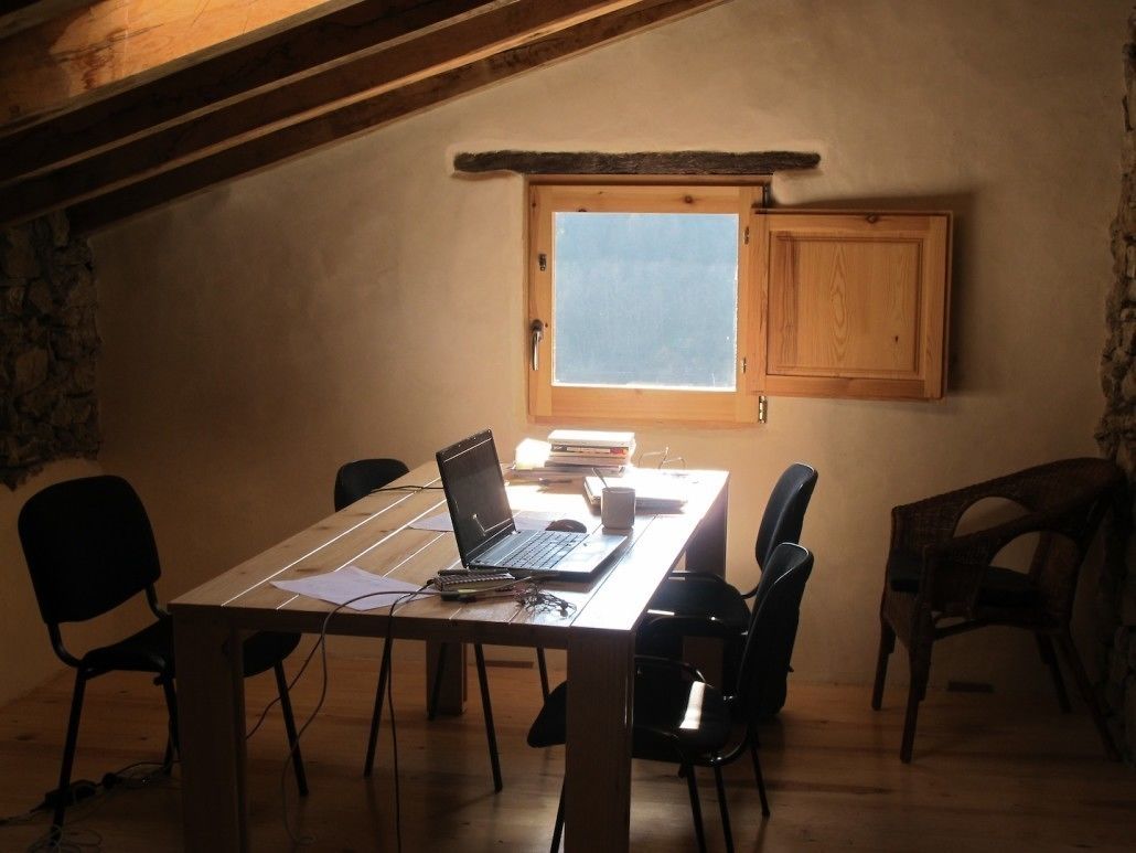 Una habitación pequeña y acogedora con una mesa de madera, una computadora portátil y sillas negras. Hay una ventana con vistas al cielo y un mueble de madera sobre la mesa.
