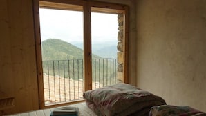 A room with a bed, a window with a view of a mountainous landscape, and a stone wall. - Alberg Rural El Negre - Hostel (Ogassa)