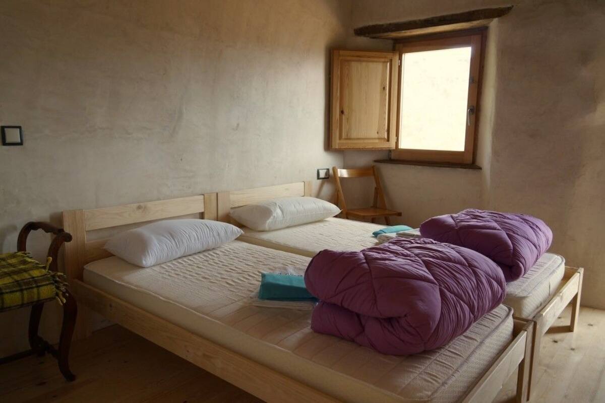 A single bed with a wooden frame, two pillows, a blanket, and a purple throw. There is a window with wooden shutters and a chair with a plaid cushion.