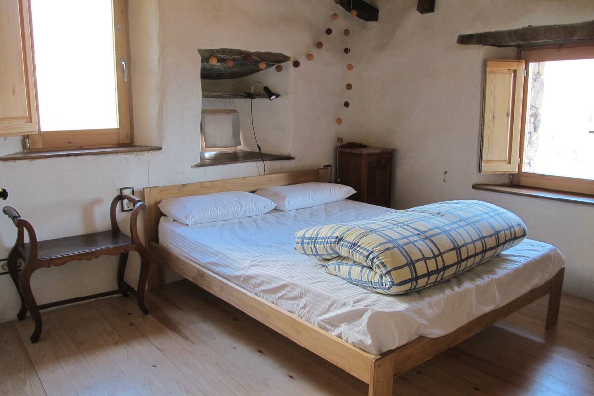 A wooden bed with white bedding and a plaid pillow, a wooden bench, and a small shelf with a lamp.