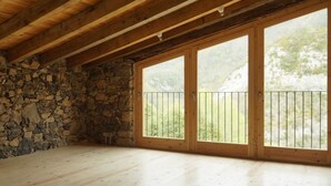 A spacious room with a stone wall, wooden floor, and large sliding glass doors with a metal railing. - Alberg Rural El Negre - Hostel (Ogassa)