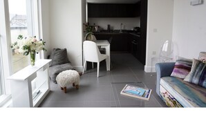 A modern living room with a sofa, a coffee table, and a dining area with a chair and table. There is a kitchen visible in the background. - Modern, Spacious 2BR Flat in Oxford (Oxford)