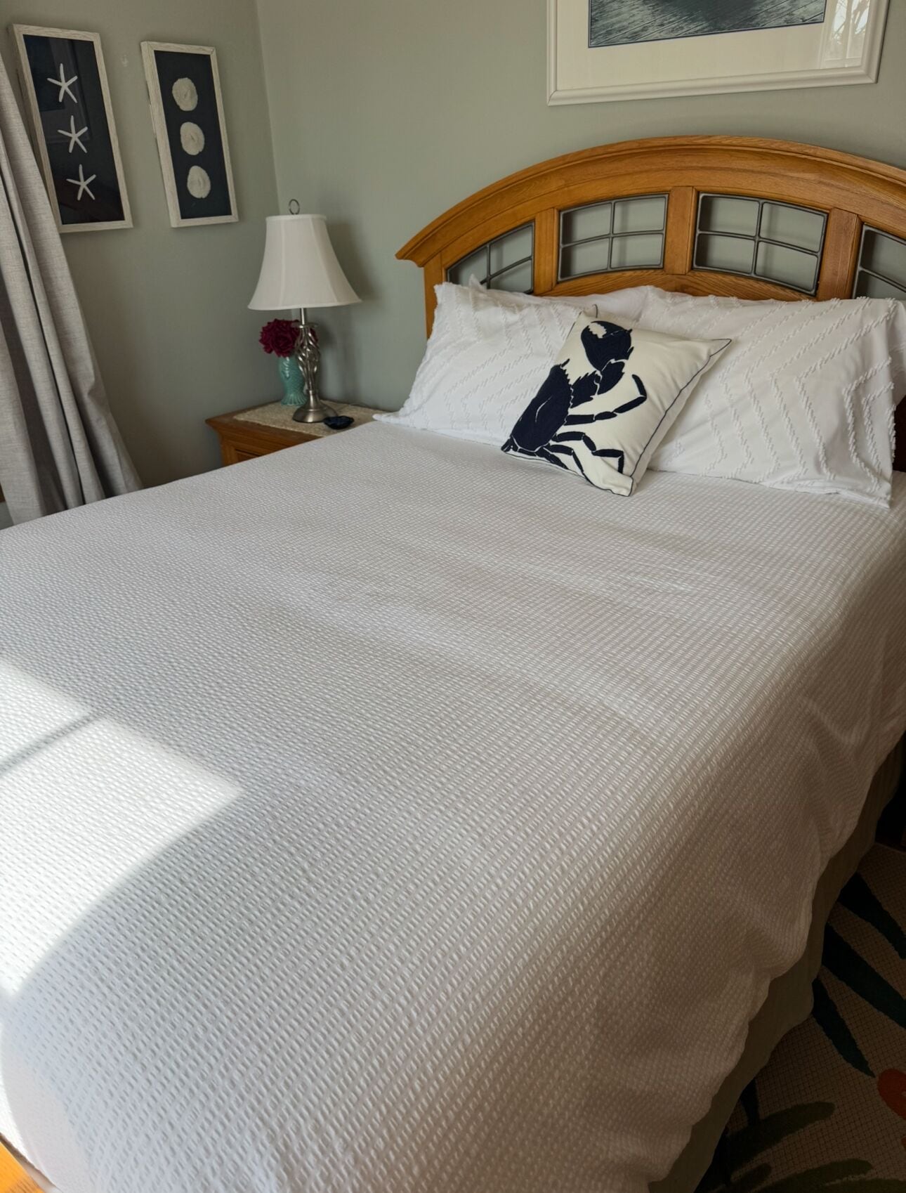 A neatly made bed with white bedding and a decorative pillow, a wooden headboard, a bedside table with a lamp, and wall art.