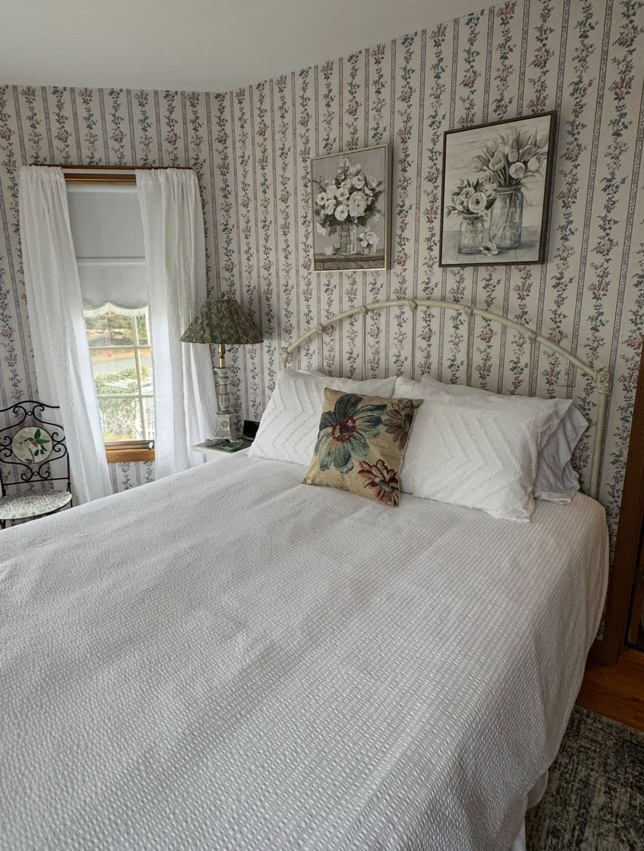 A bedroom with a large bed, floral wallpaper, a window with curtains, and two framed floral paintings on the wall.