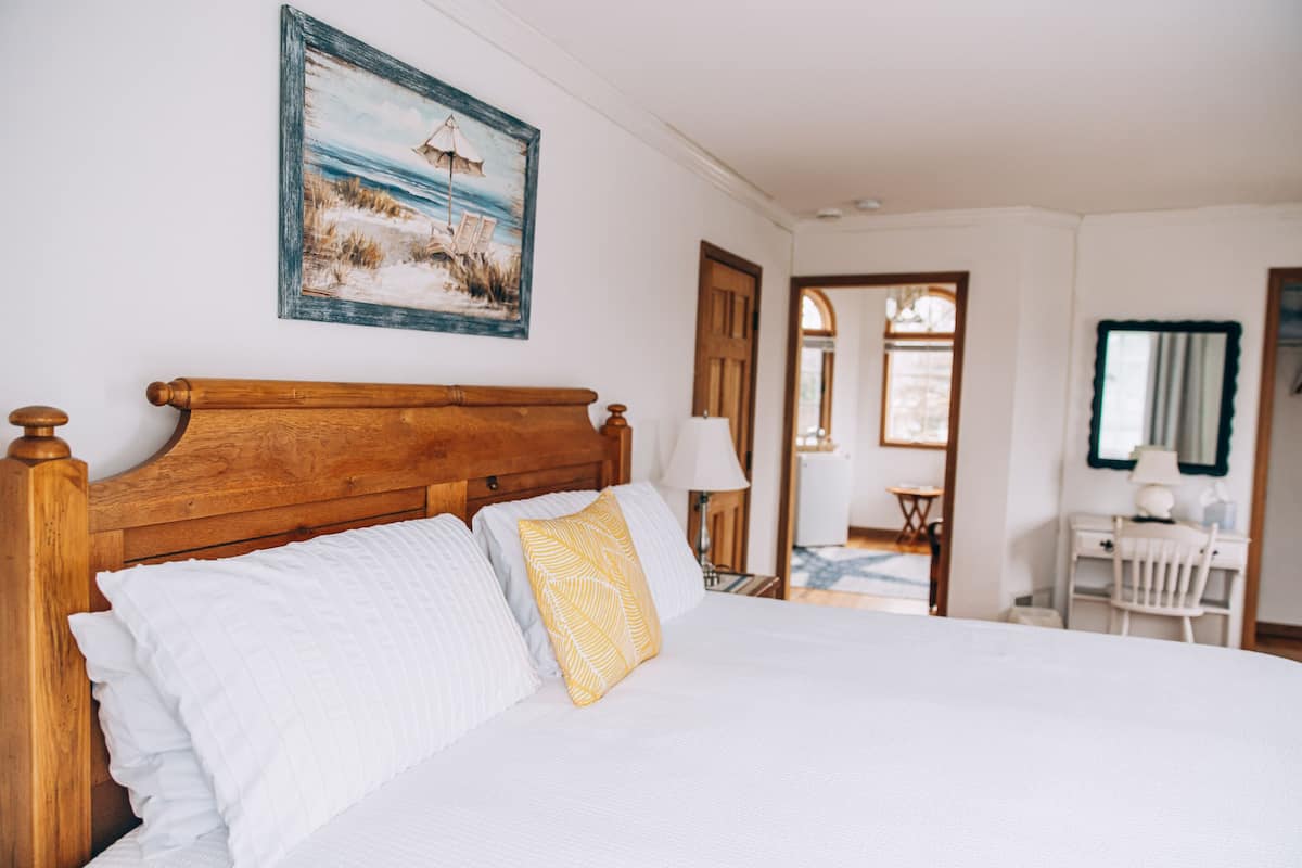 Uma cama bem arrumada com cabeceira de madeira, almofadas brancas e estampadas, e um quadro com uma paisagem de praia acima.