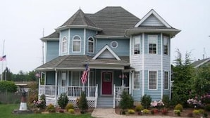 A two-story house with a porch, a red door, and a small garden with flowers and shrubs. - Captain's Cottage (Forked River)