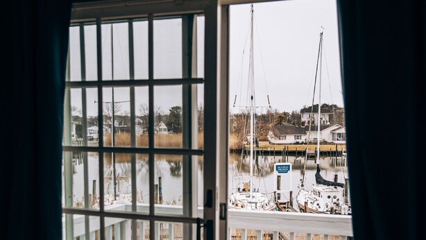 A room with a large sliding glass door leading to a wooden deck with a view of a marina and boats. - Captain's Cottage (Forked River)