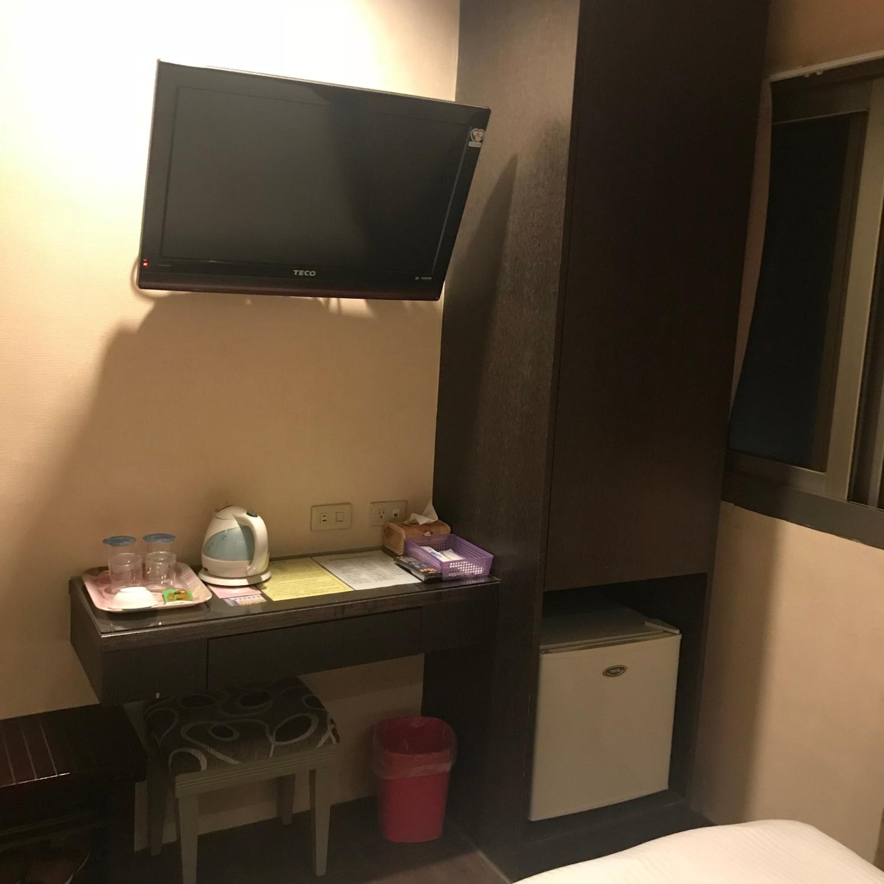 Ein Hotelzimmer mit an der Wand befestigtem Fernseher, einem kleinen Schreibtisch mit Wasserkocher, Tassen und einem Korb, einer Minibar und einem Mülleimer.