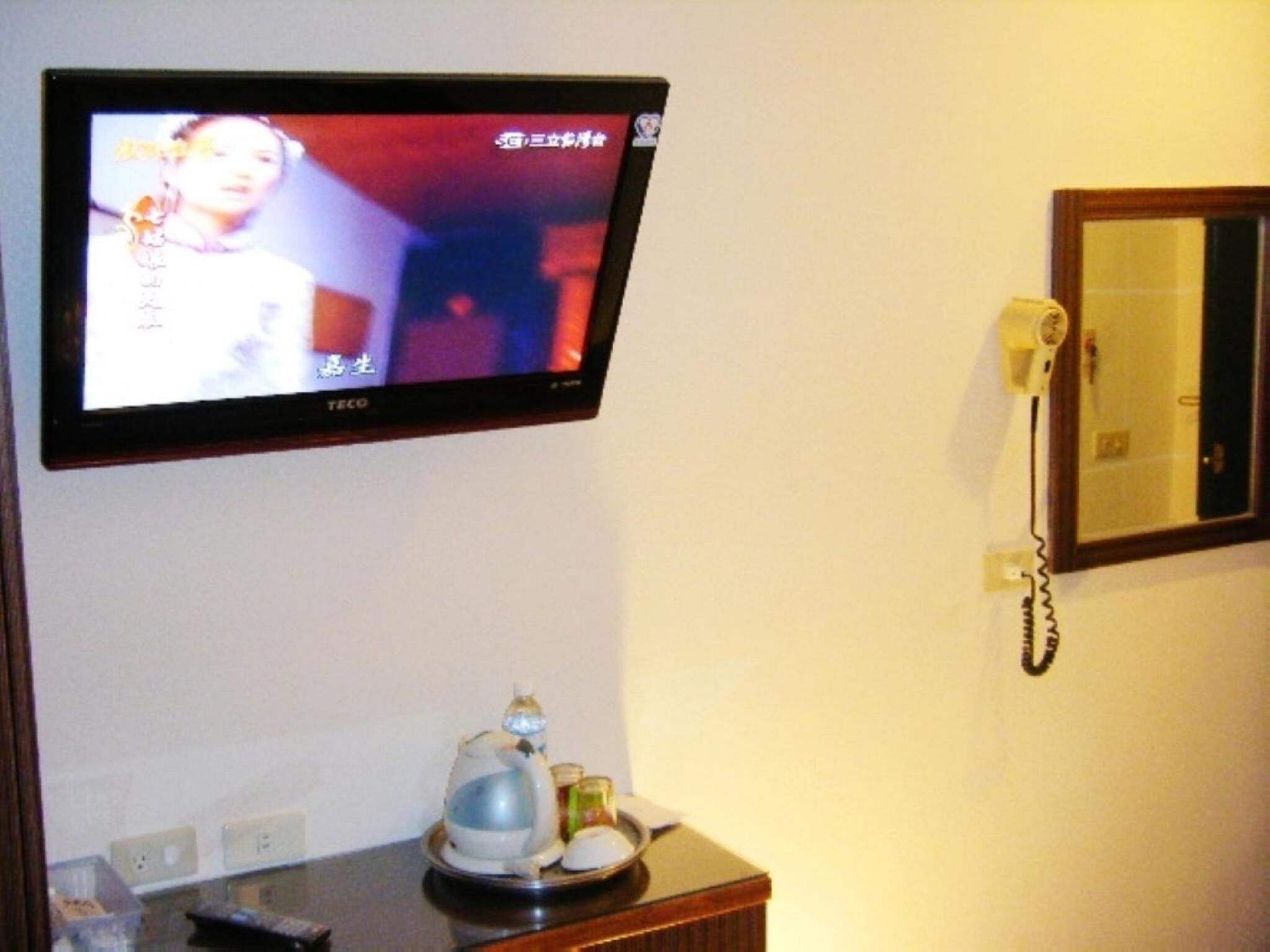 A television mounted on a wall displaying a program in a foreign language. Below the TV, there is a small table with a kettle, a cup, and a remote control. A telephone is mounted on the wall to the right of the TV.