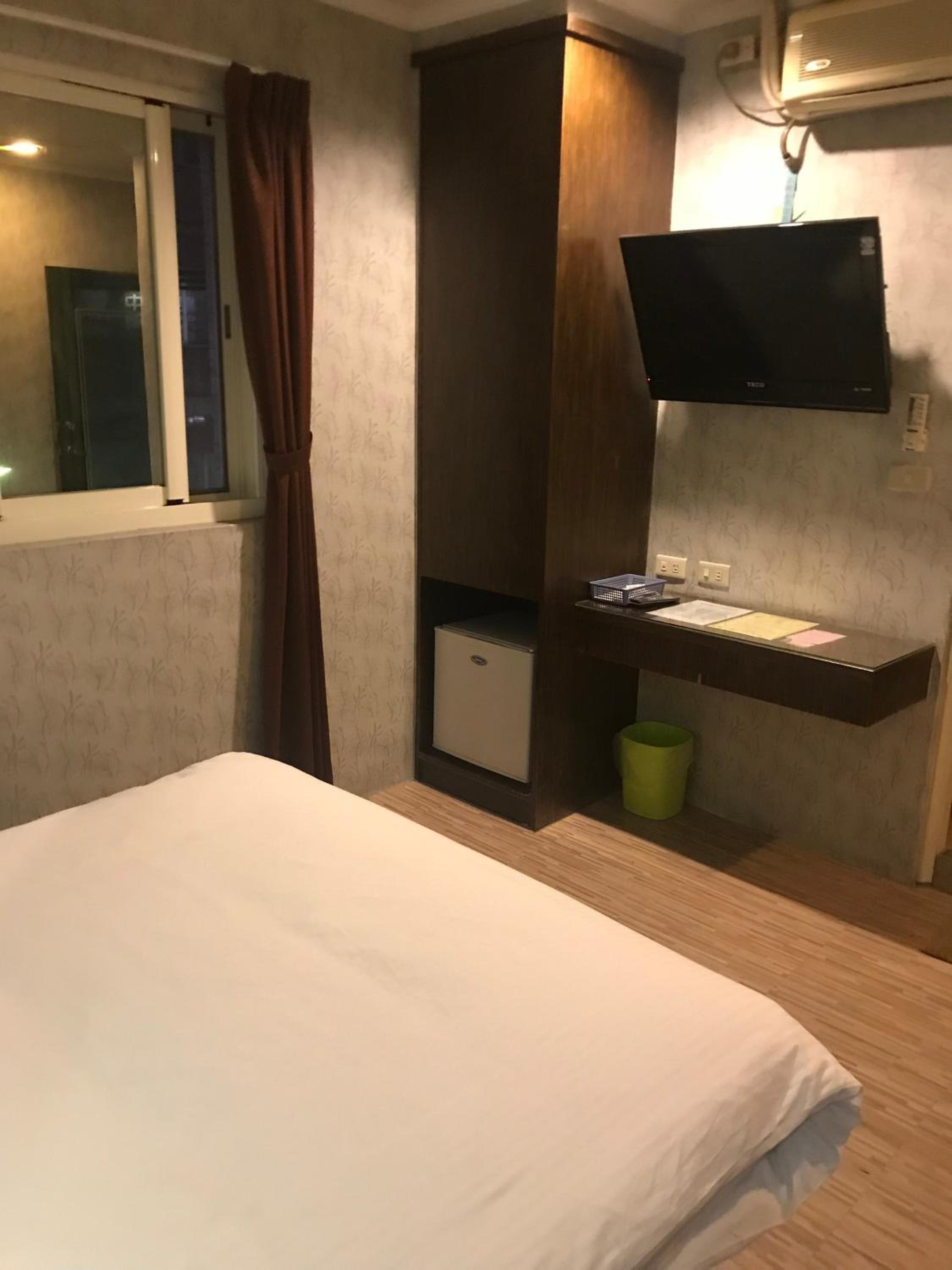 A hotel room with a bed, a small desk with a computer, a TV mounted on the wall, and a window with curtains.