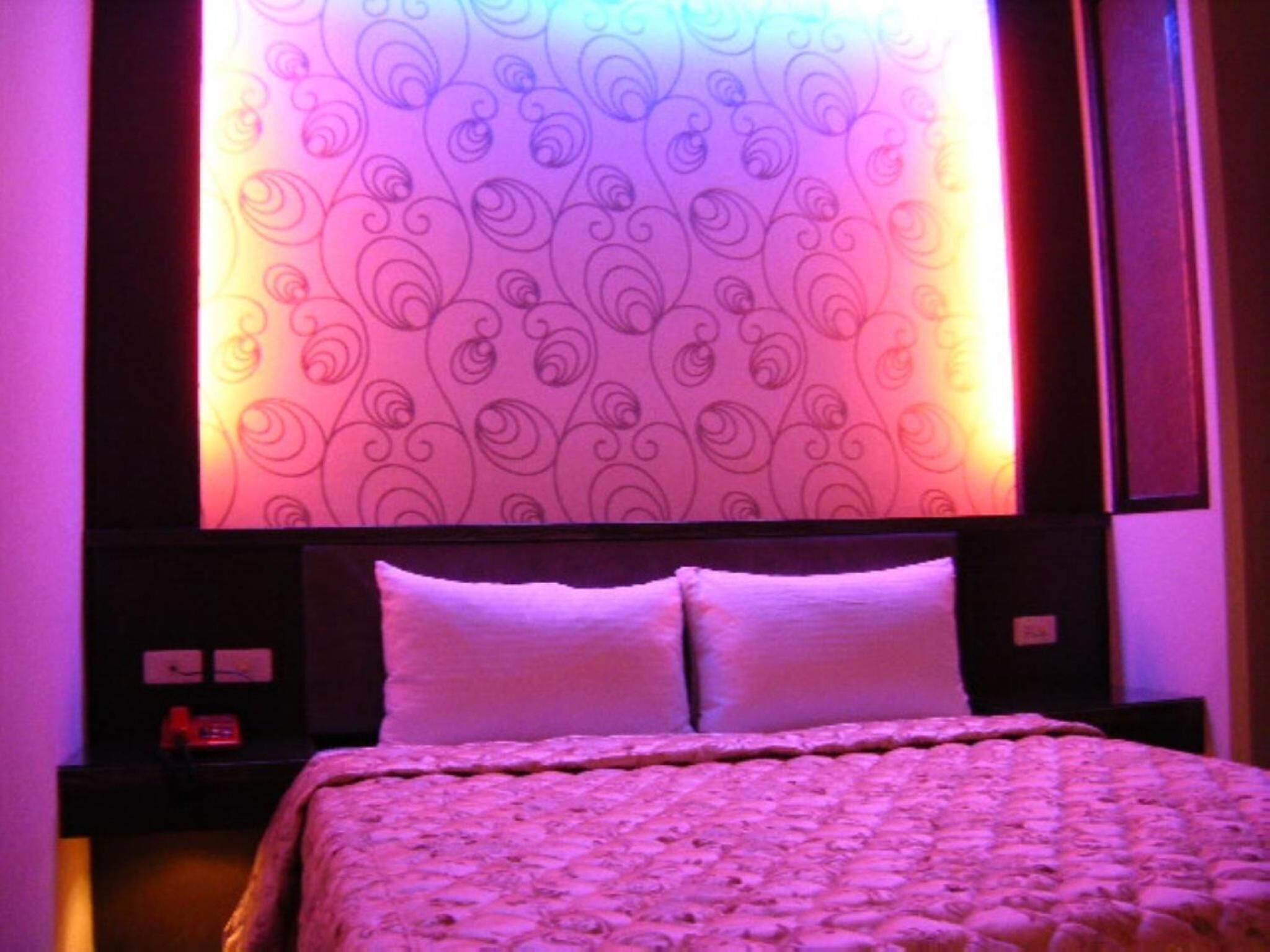 A bed with a patterned headboard and two pillows, a nightstand with a clock, and a wall with a decorative wallpaper.