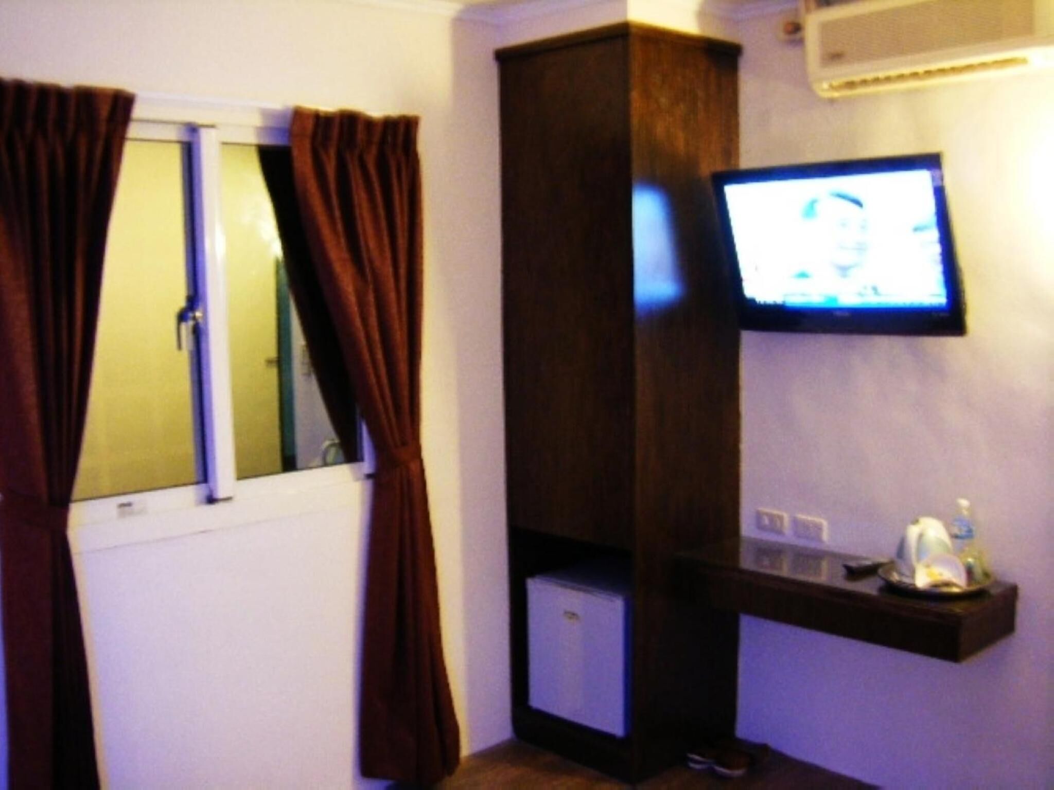 A hotel room with a television mounted on the wall, a small cabinet below it, and a window with curtains.