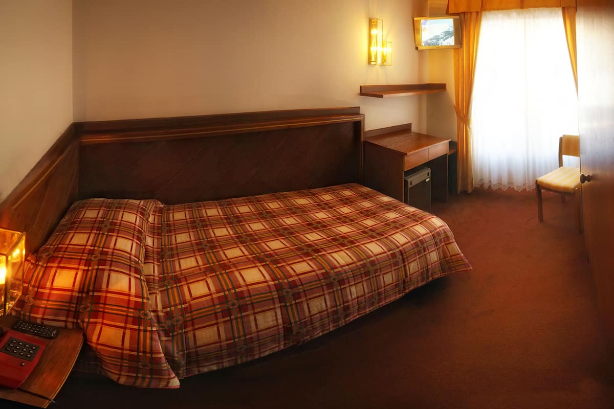 Habitación de hotel con una cama individual, mesita de noche, lámpara y un pequeño escritorio con una silla.
