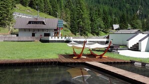 Un espace piscine avec une terrasse en bois, un bâtiment doté d’un toit en pente et un paysage montagneux en arrière-plan.