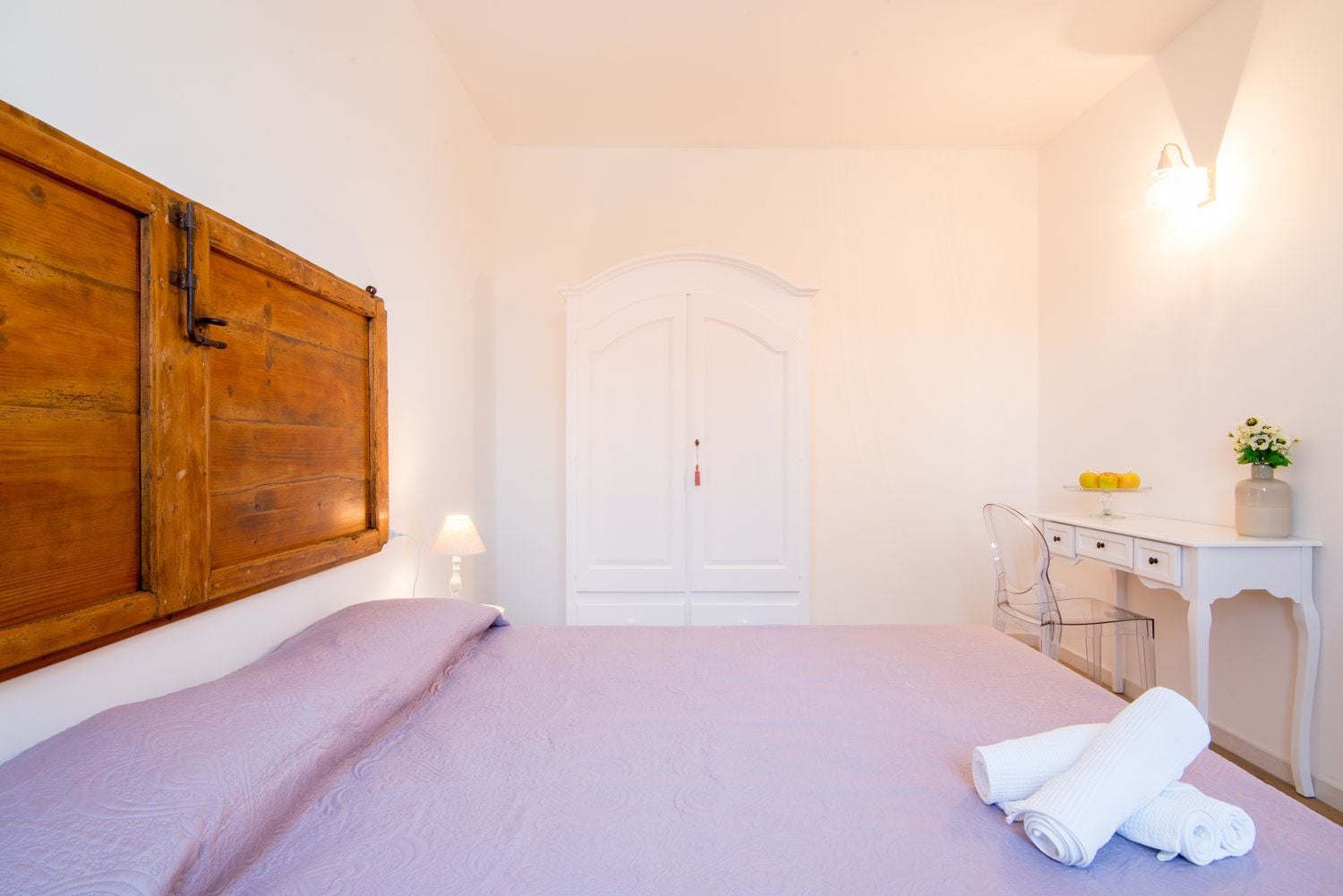 A bedroom with a bed, a wooden wardrobe, a desk with a chair, and a white door.