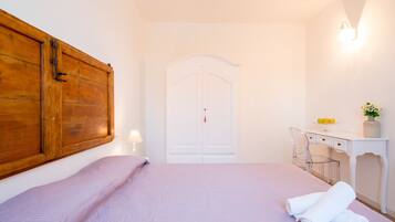 A bedroom with a bed, a wooden wardrobe, a desk with a chair, and a white door.