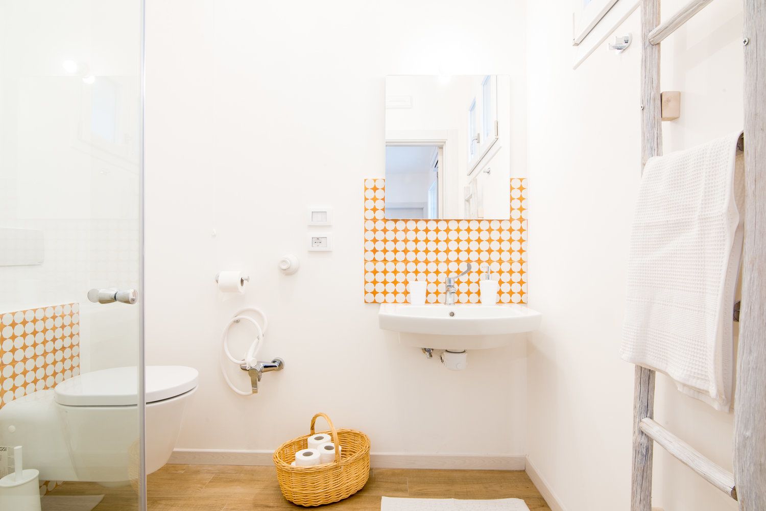 A bathroom with a toilet, sink, and mirror. There is a basket with toilet paper on the floor.