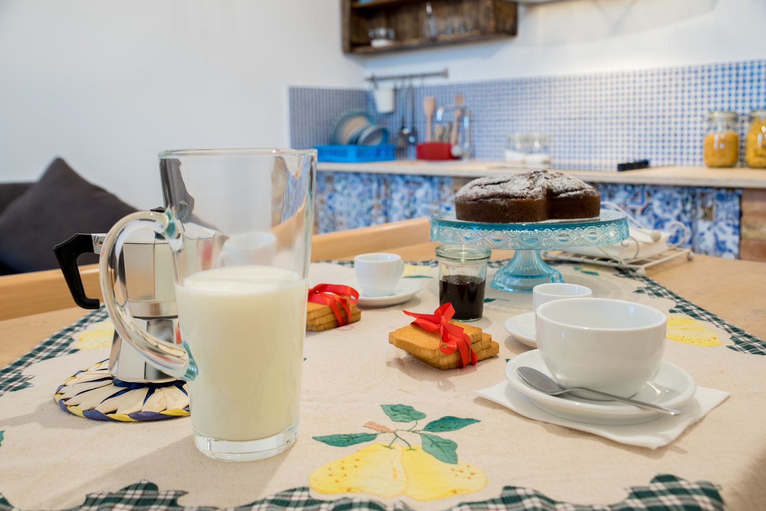 Una mesa con mantel floral, con un jarro de leche, tazas, un pastel y galletas.