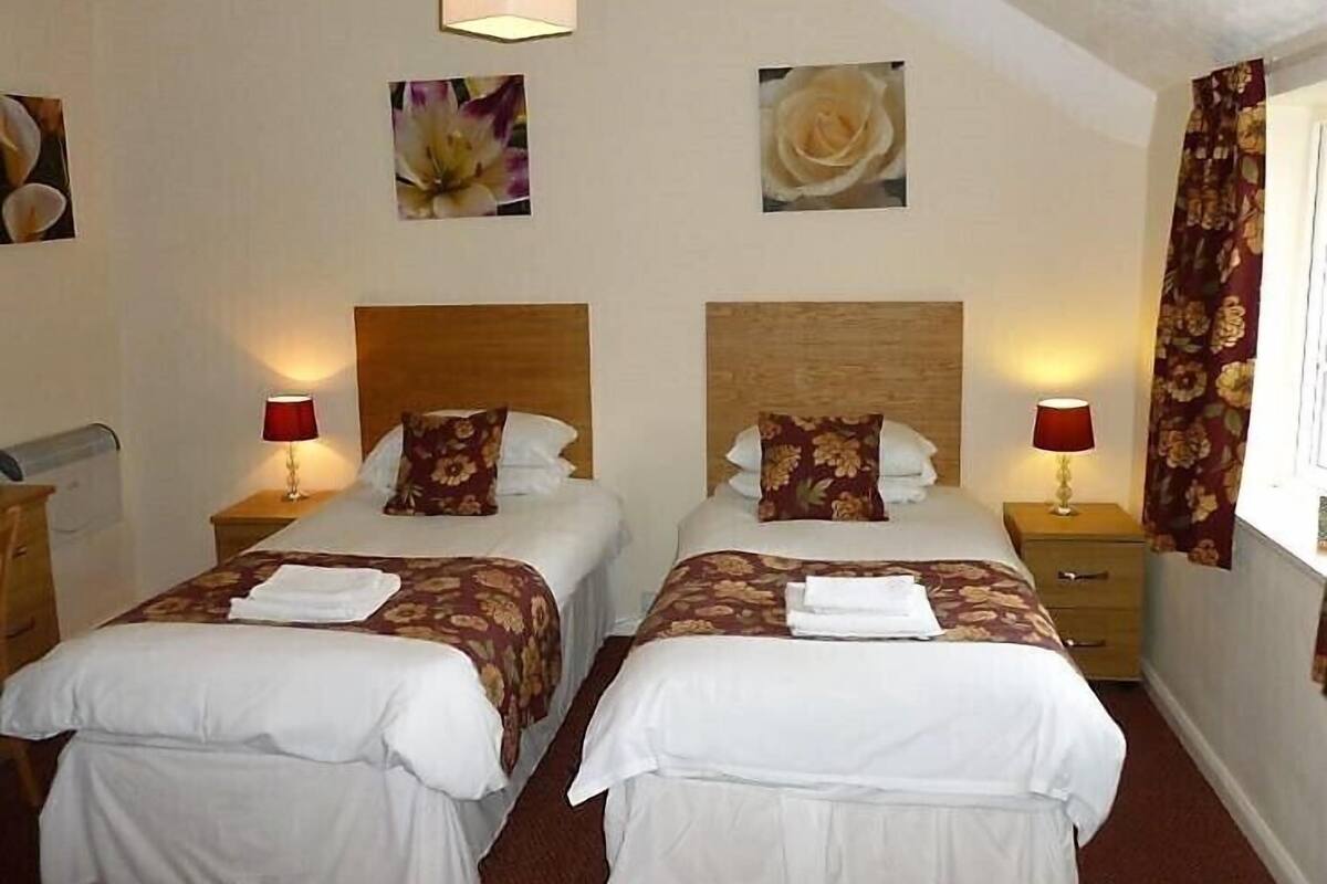 A hotel room with two beds, each with a wooden headboard, white bedding, and floral-patterned pillows. There are two bedside tables with lamps, a window with curtains, and wall art depicting flowers.