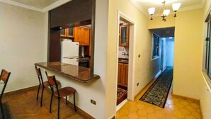Une cuisine compacte équipée d’un réfrigérateur, d’une cuisinière et de meubles de rangement en bois. On y trouve également un coin repas avec une table et des chaises. Un couloir, agrémenté d’un tapis à motifs, mène à une porte.