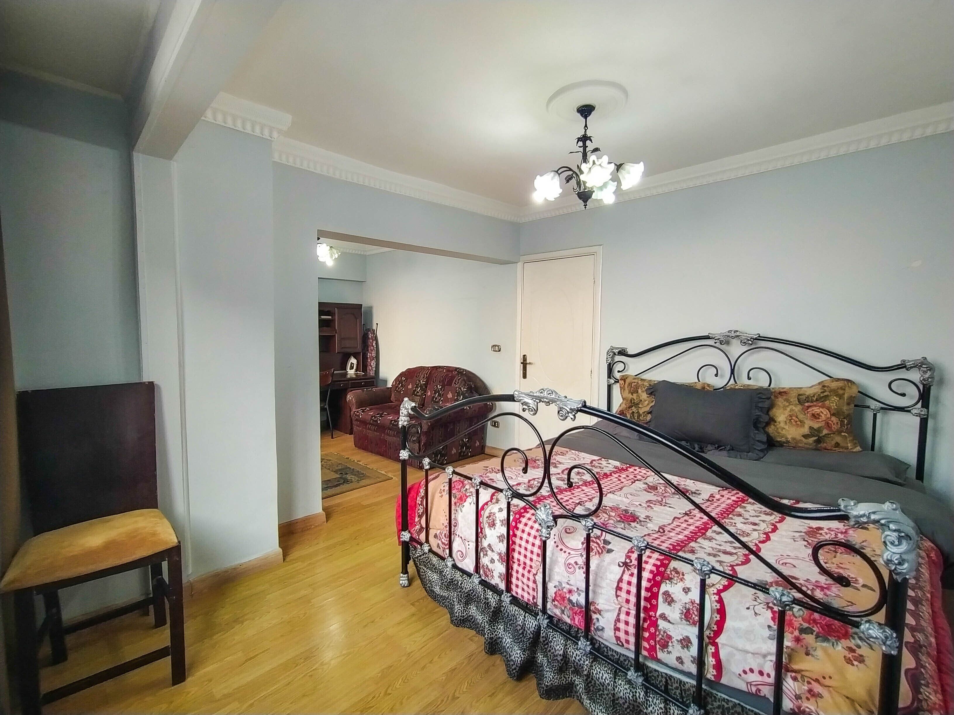 Un dormitorio con cama, una silla, suelo de madera, luz en el techo y una puerta que da a otra habitación.