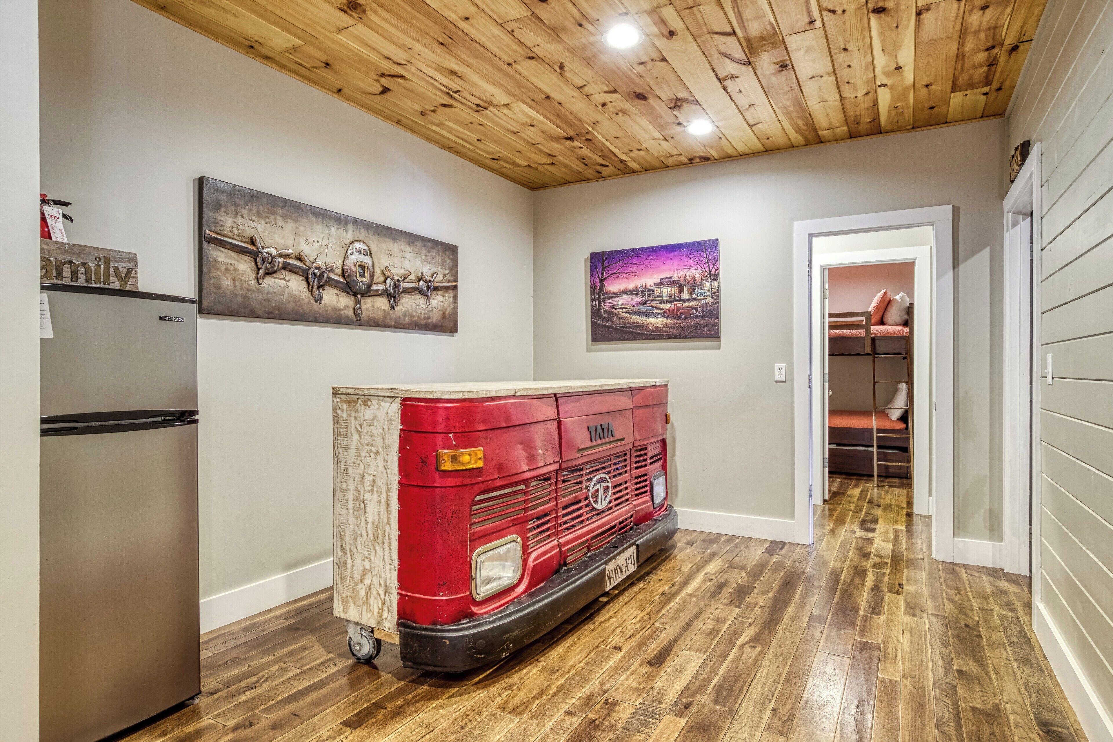 A hallway with a wooden ceiling, a red vintage van, a refrigerator, and two wall-mounted paintings.