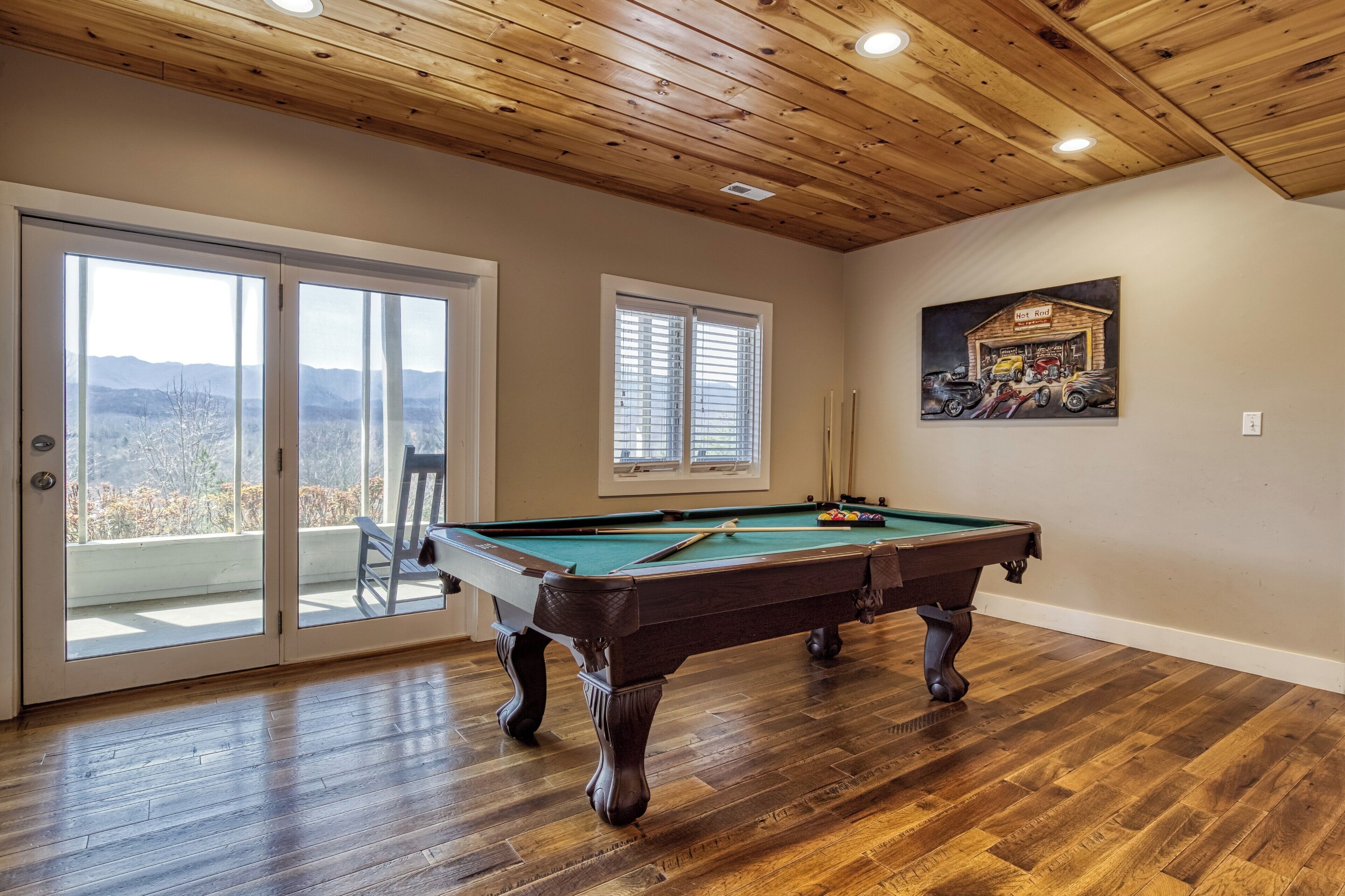 A pool table in a room with wooden flooring and a wooden ceiling, large windows with blinds, and a painting on the wall.