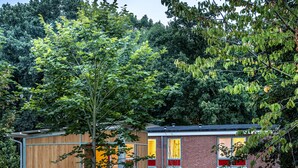 Un edificio moderno con fachada de madera y ventanas de vidrio, rodeado de abundante vegetación y mobiliario exterior.