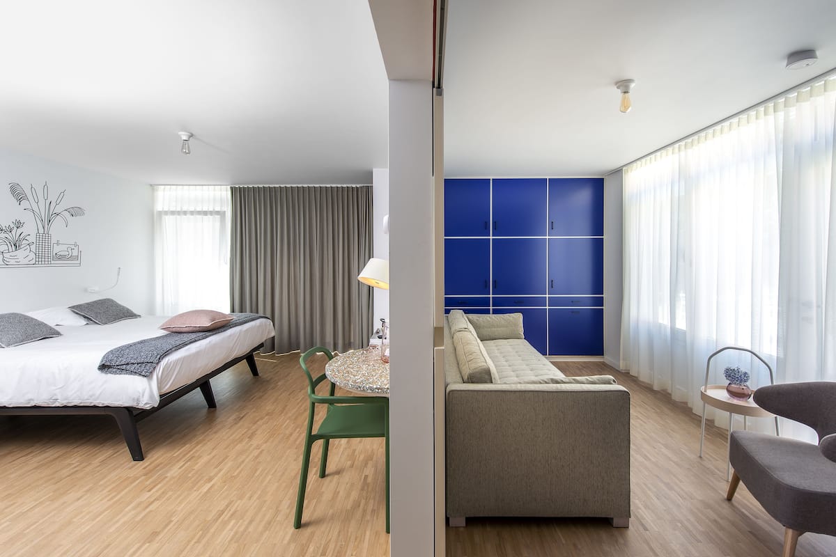 A modern hotel room with a bed, a sofa, a dining table, and a blue wardrobe.