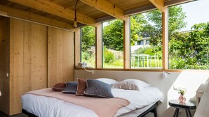 Un dormitorio con cama, paredes de madera, una ventana que da a zonas verdes y una mesita con un florero.