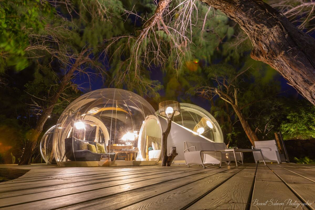 A transparent dome structure with seating and a table, surrounded by trees and a wooden deck.